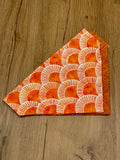 A triangular orange and sunrise patterned dog bandana laid flat on a wooden surface.