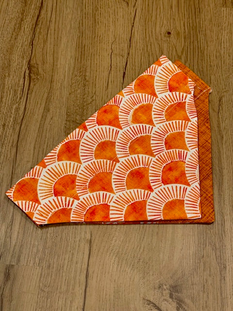 A triangular orange and sunrise patterned dog bandana laid flat on a wooden surface.