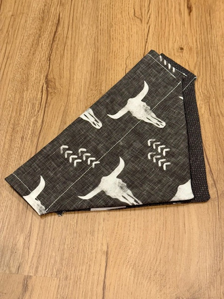 A black and white dog bandana with a cow skull print, displayed on a wooden floor.