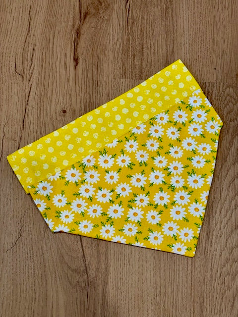 A yellow dog bandana with white daisies and polka dots pattern laid out flat on a wooden surface.