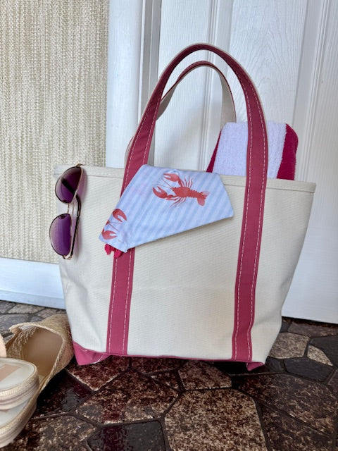 Beige tote bag with lobster dog bandana, sunglasses, and a towel on a textured surface.